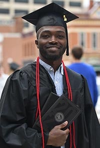 A Western graduate smiles as he holds his diploma in his cap and gown.