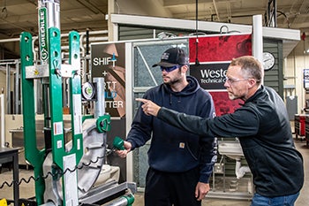 An instructor shows an apprentice how to operate a machine in a workshop.
