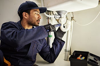 A plumbing apprentice inspects a pipes beneath an industrial sink.