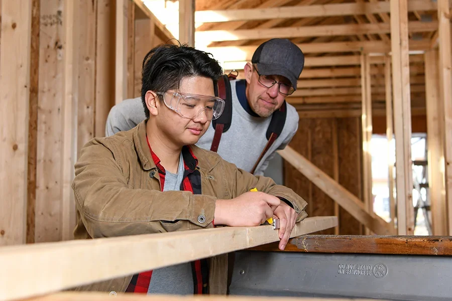 An instructor supervises a student as he measures lumber for a building.