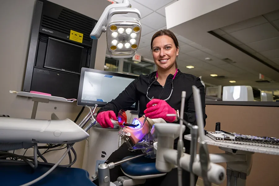 A dental assistant student poses with equipment in a dentist's exam room.