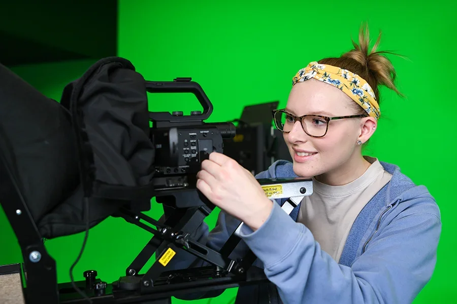 A female student operates a television camera in front of a green screen.