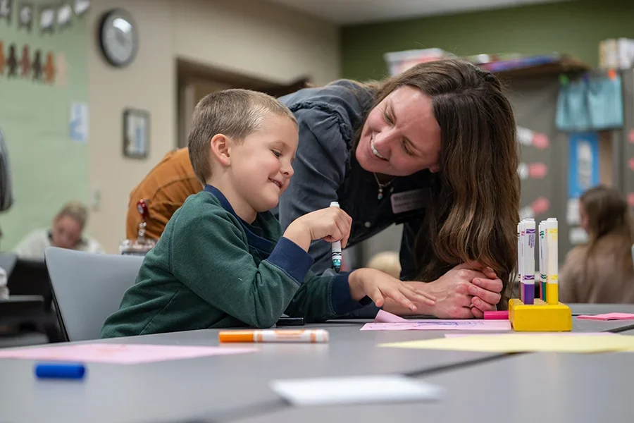 An education students works with a young boy at his desk in a classroom.