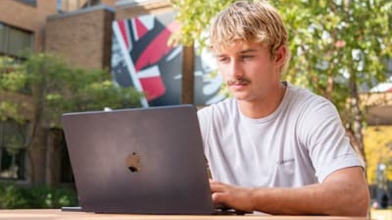 A student sits at a picnic table working on his laptop.