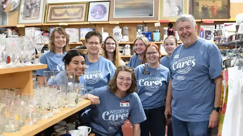 A group of smiling people wearing matching blue "Western Cares" T-shirts poses in a thrift store, surrounded by shelves of glassware and mugs.