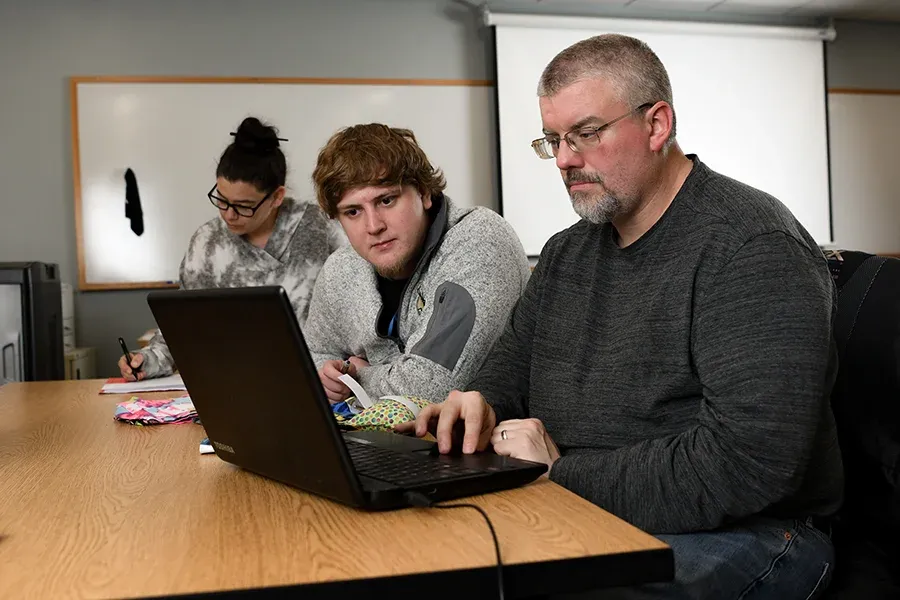 An accounting instructor works with a student on a laptop in a classroom.