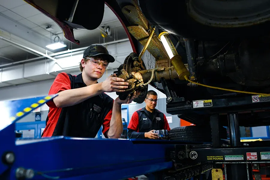 Students work on the underside of vehicles in an auto shop.