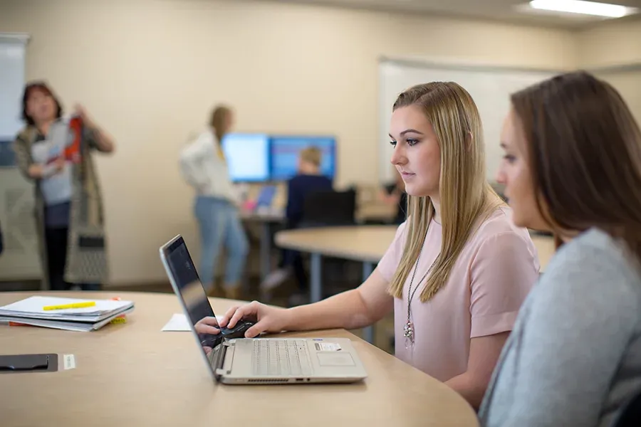 Two students sit at a laptop and collaborate in a classroom.