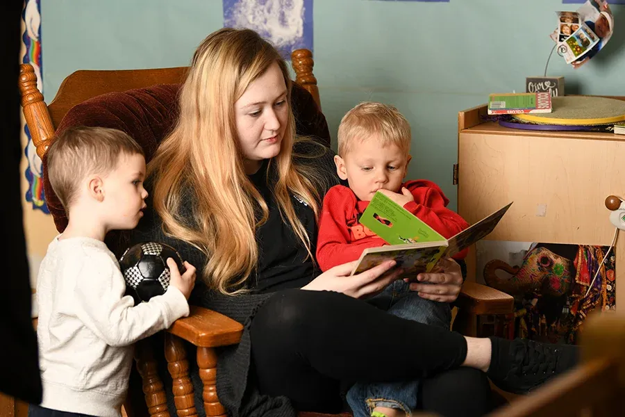 A student reads to two young boys in a rocking chair in a classroom.