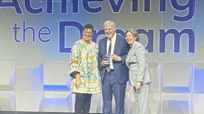 Three people stand on a stage holding an award with "Achieving the Dream" text behind them.