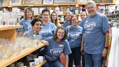 A group of smiling people wearing matching blue "Western Cares" T-shirts poses in a thrift store, surrounded by shelves of glassware and mugs.