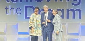 Three people stand on a stage holding an award with "Achieving the Dream" text behind them.