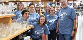 A group of smiling people wearing matching blue "Western Cares" T-shirts poses in a thrift store, surrounded by shelves of glassware and mugs.