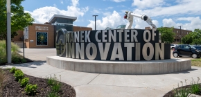 Entrance of Wanek Center of Innovation with metallic signage and robotic arm sculpture in front of a modern building.